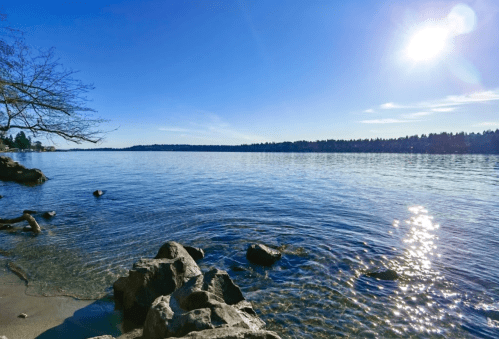 A serene lakeside view with clear water, rocky shore, and a bright sun shining in a blue sky.