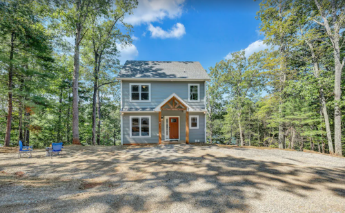 A modern two-story house surrounded by trees, with a gravel driveway and two chairs in front.