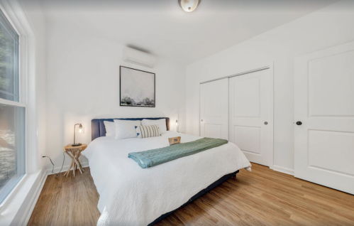 Bright, minimalist bedroom with a large bed, wooden floor, and natural light from a window.