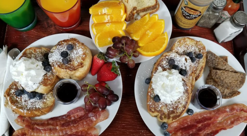 Two plates of breakfast featuring French toast with blueberries, whipped cream, bacon, fruit, and drinks in colorful glasses.