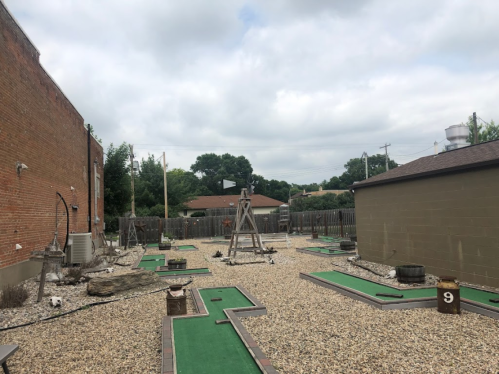 A mini-golf course with green turf and gravel, surrounded by buildings and trees under a cloudy sky.