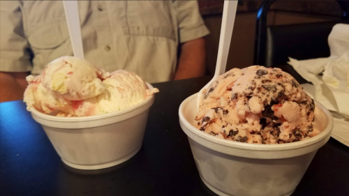 Two cups of ice cream on a table: one with strawberry and vanilla, the other with chocolate and pink swirls.