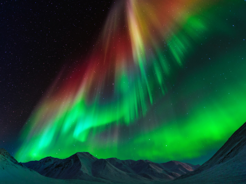 Vibrant auroras dance across the night sky, illuminating snowy mountains with shades of green, red, and purple.