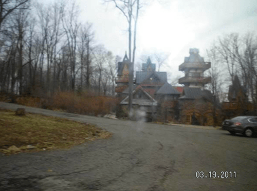A winding road leads to a large, ornate mansion surrounded by bare trees on a cloudy day.