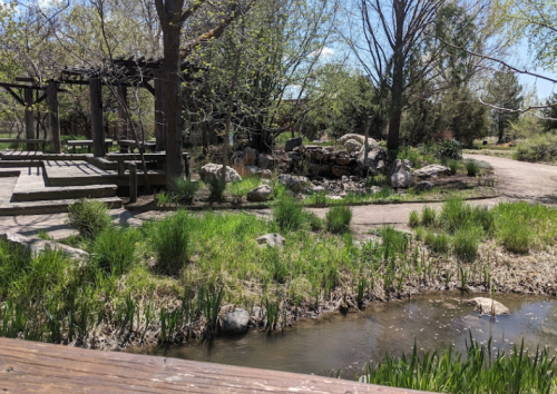 A serene park scene featuring a stream, grassy areas, and a wooden gazebo surrounded by trees.