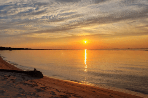 A serene sunset over calm waters, with soft clouds and a sandy beach in the foreground.