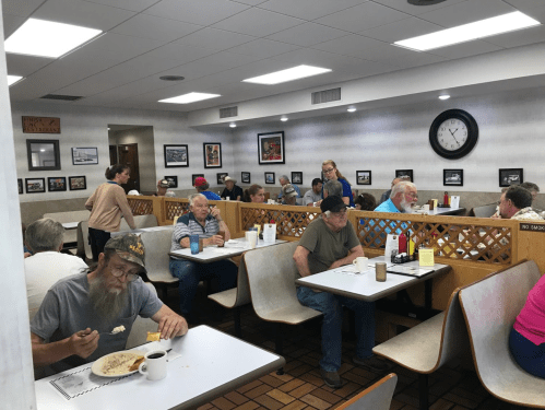 A busy diner with patrons eating at booths, a waitress serving, and a clock on the wall.