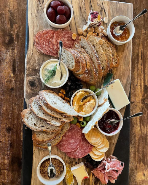 A wooden platter filled with assorted breads, meats, cheeses, olives, nuts, and small bowls of spreads and jams.
