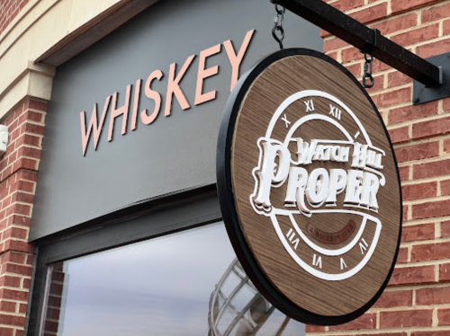 Sign for "Watch Hill Proper" with a whiskey theme, hanging from a brick building.