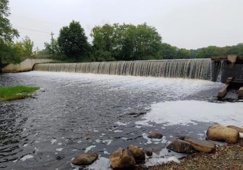 A serene view of a waterfall cascading over a dam, surrounded by greenery and rocky shoreline.