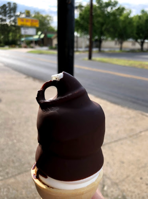 A hand holding a chocolate-dipped ice cream cone with a bite taken out, set against a street scene.