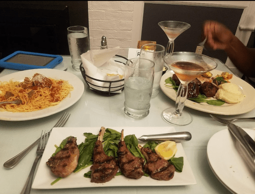 A dining table with spaghetti, lamb chops on spinach, and cocktails, set in a cozy restaurant atmosphere.
