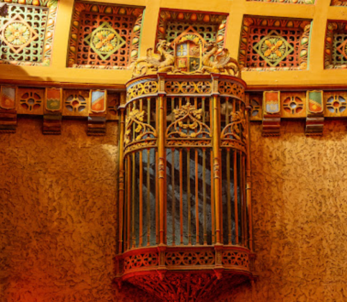 Ornate wooden balcony with intricate carvings, set against a textured wall and colorful ceiling details.