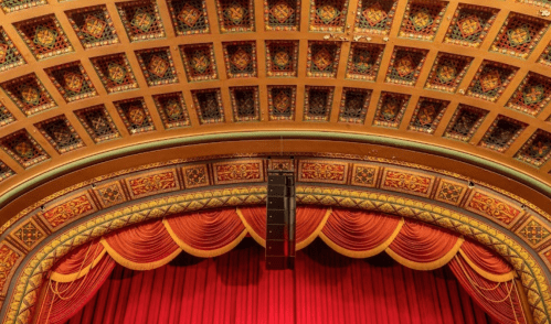 Ornate theater ceiling with intricate patterns and a red curtain below, showcasing a grand architectural design.