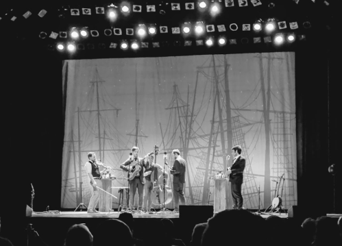 A black and white photo of a band performing on stage with a nautical-themed backdrop and audience in the foreground.