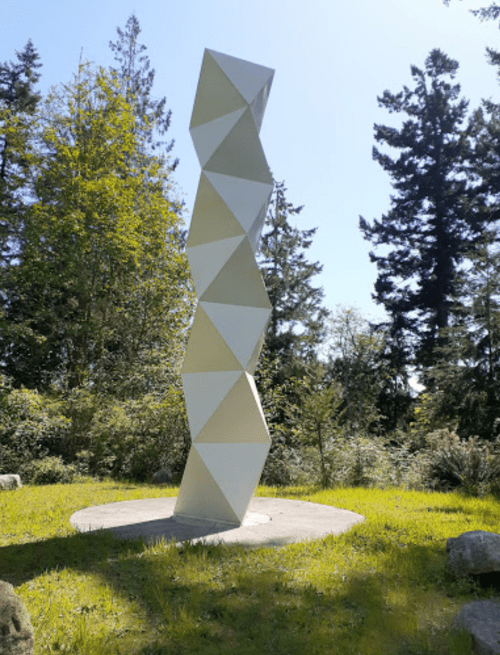 A tall, geometric sculpture made of white panels stands in a grassy area surrounded by trees.