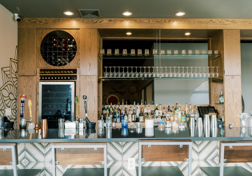 A stylish bar with a wooden counter, shelves of glassware, and a variety of bottles displayed behind the counter.