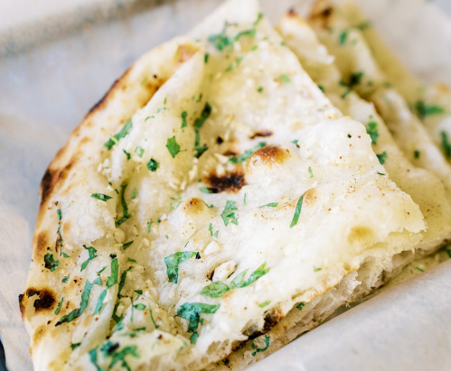 A close-up of a piece of garlic naan topped with chopped herbs, resting on parchment paper.