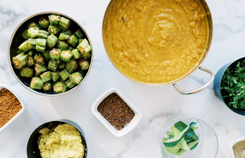 A top-down view of a kitchen counter with bowls of okra, lentil soup, spices, and fresh herbs.