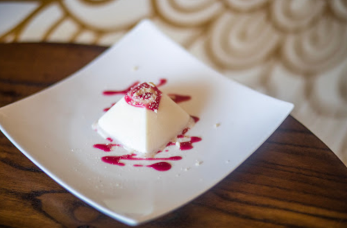 A creamy dessert shaped like a pyramid, topped with a strawberry and drizzled with raspberry sauce on a white plate.
