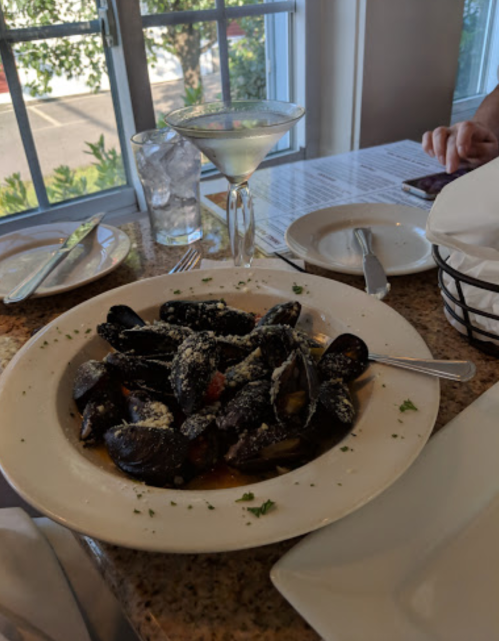 A plate of mussels in a white sauce, garnished with herbs, next to a martini on a table by a window.