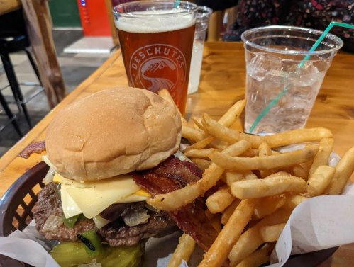 A burger with bacon and cheese, served with fries, alongside a pint of beer and a cup of water on a wooden table.