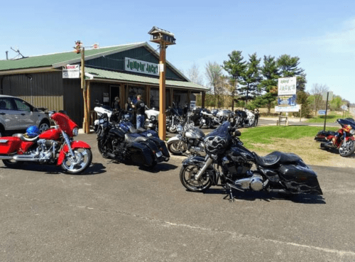 A row of motorcycles parked outside a shop called "Jumper Jack's" on a sunny day. Trees in the background.
