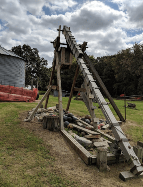 A wooden catapult structure with a ramp, set in a grassy area near a barn, surrounded by rocks and a safety barrier.