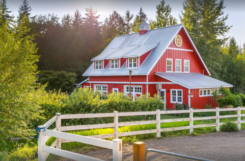 A charming red barn-style house surrounded by greenery and a white fence, set against a backdrop of trees.