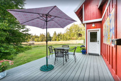 A red house with a patio featuring a table, chairs, and a large umbrella, surrounded by greenery and open fields.