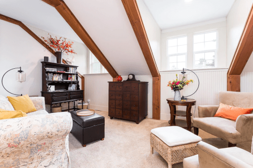 Cozy attic living space with wooden beams, a bookshelf, armchairs, and decorative flowers. Soft lighting enhances warmth.