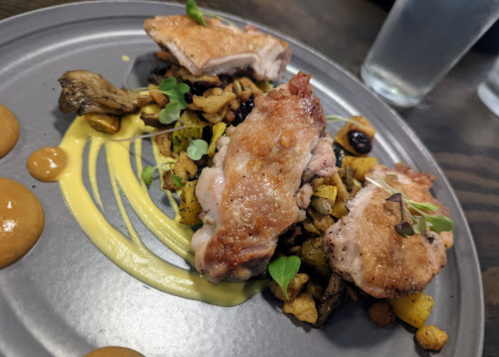 Plated dish featuring seared chicken, roasted vegetables, and sauces on a gray plate, with water glasses in the background.