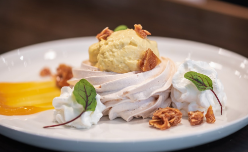 A dessert featuring meringue, creamy yellow ice cream, whipped cream, and garnished with crispy bits and green leaves.