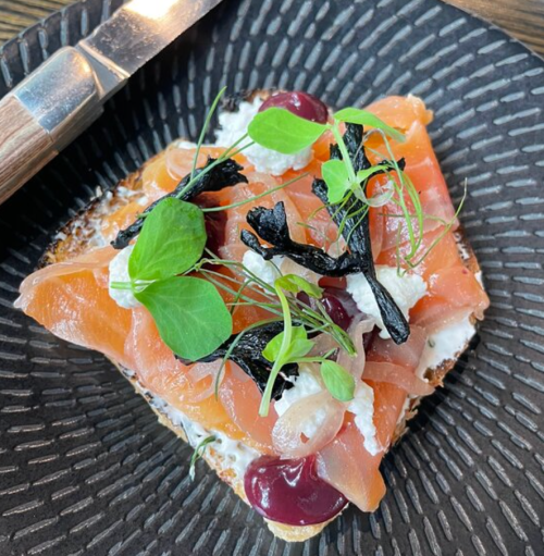 A slice of bread topped with smoked salmon, cream cheese, black seaweed, and garnished with microgreens on a textured plate.