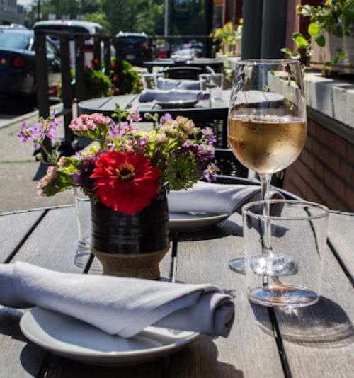 A table set for dining outdoors, featuring a glass of white wine, a flower arrangement, and neatly folded napkins.
