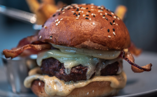 A close-up of a juicy burger with melted cheese, bacon, and fries on the side, served on a sesame seed bun.