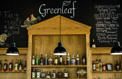 A wooden shelf with various bottles, under a chalkboard featuring "Greenleaf" and farm-related illustrations.