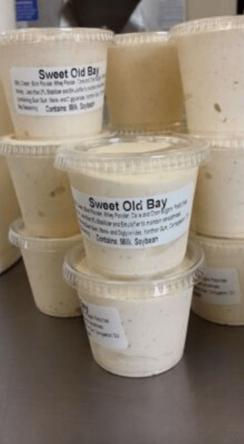 Containers of "Sweet Old Bay" sauce stacked on a countertop, labeled with ingredients and allergen information.