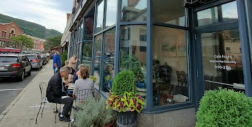 A café with outdoor seating, people dining, and a view of a street lined with shops and mountains in the background.