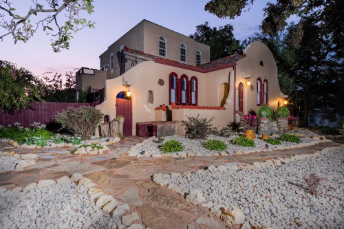 A charming adobe-style house with colorful windows, surrounded by a landscaped garden and stone pathway at dusk.