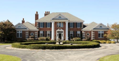 A large brick house with a circular driveway, manicured lawns, and multiple chimneys under a clear blue sky.