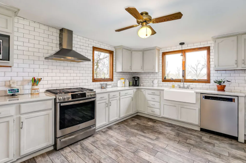 Bright kitchen with white cabinets, stainless steel appliances, and a farmhouse sink, featuring large windows and wood flooring.