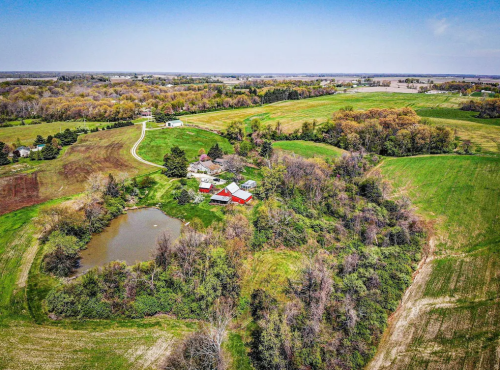 Aerial view of a rural landscape featuring a pond, fields, and a cluster of buildings surrounded by trees.