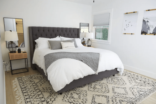 A modern bedroom featuring a gray tufted bed, white bedding, two lamps, and a patterned rug.