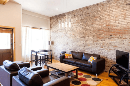Modern living room with exposed brick wall, leather sofas, a wooden coffee table, and a dining area with a small table.