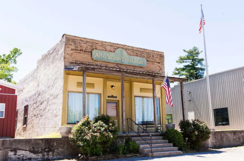 A rustic building with a sign reading "Alto Wine Depot" and American flags, surrounded by greenery and other structures.
