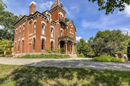 A large, red brick Victorian-style house surrounded by greenery and a gravel path under a blue sky.
