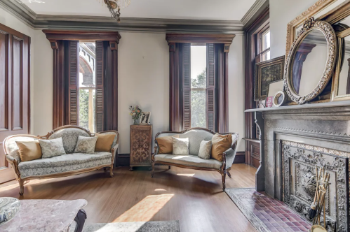 Bright living room with vintage sofas, wooden shutters, and a decorative fireplace. Natural light fills the space.