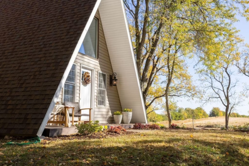 A cozy A-frame house surrounded by trees and autumn foliage, featuring a welcoming porch and decorative planters.