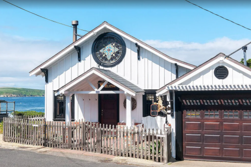 A charming white house with a circular clock, surrounded by a picket fence and near the ocean.